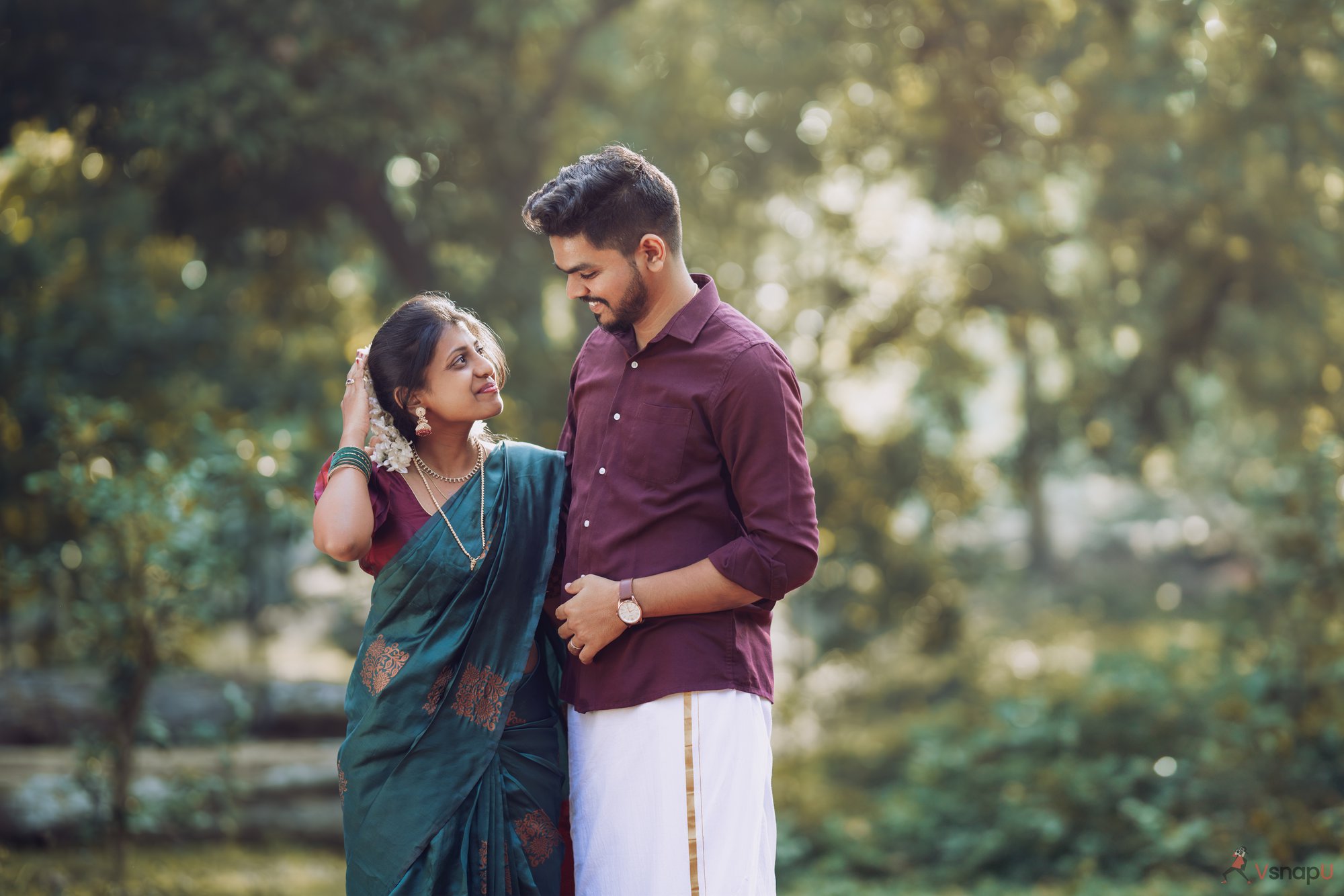 South Indian couple standing in a lush landscape, bride tenderly playing with the groom's hair while he admires her.
